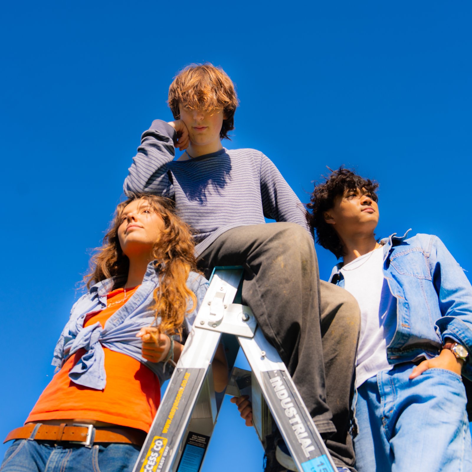 Three members of Juna shot from below against a bright blue sky, one sitting on top of a stepladder